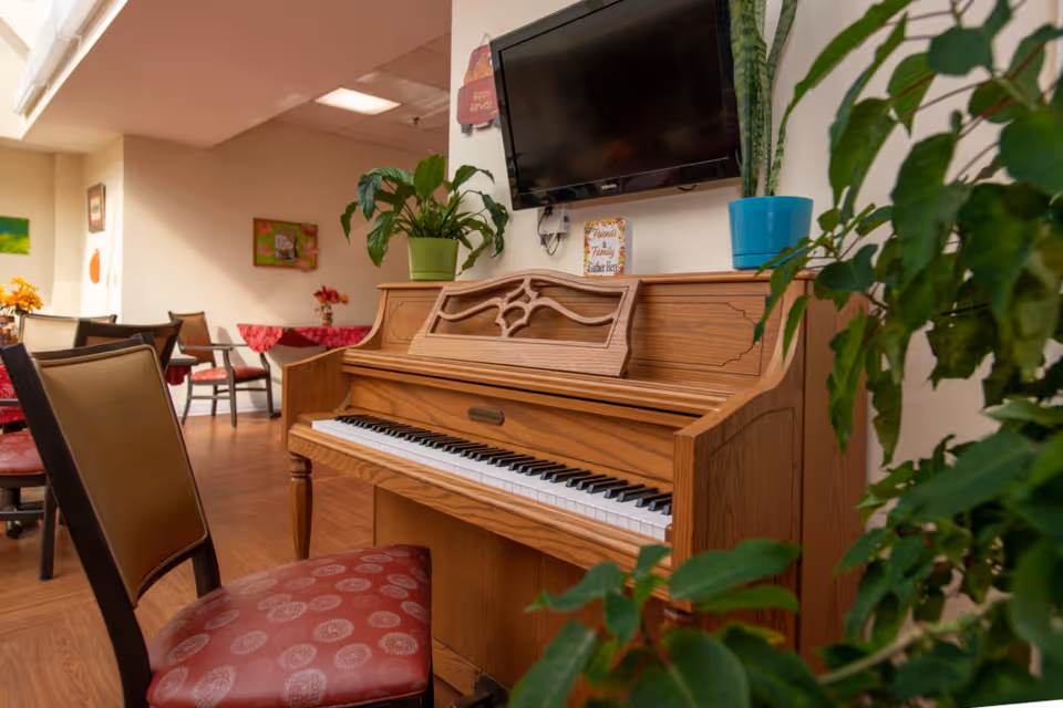 A cozy common area featuring a wooden upright piano with a decorative music stand, a flat-screen TV mounted on the wall above it, and several potted plants placed on and around the piano. In the background, there are tables and chairs with red patterned cushions, wall decorations, and warm lighting.