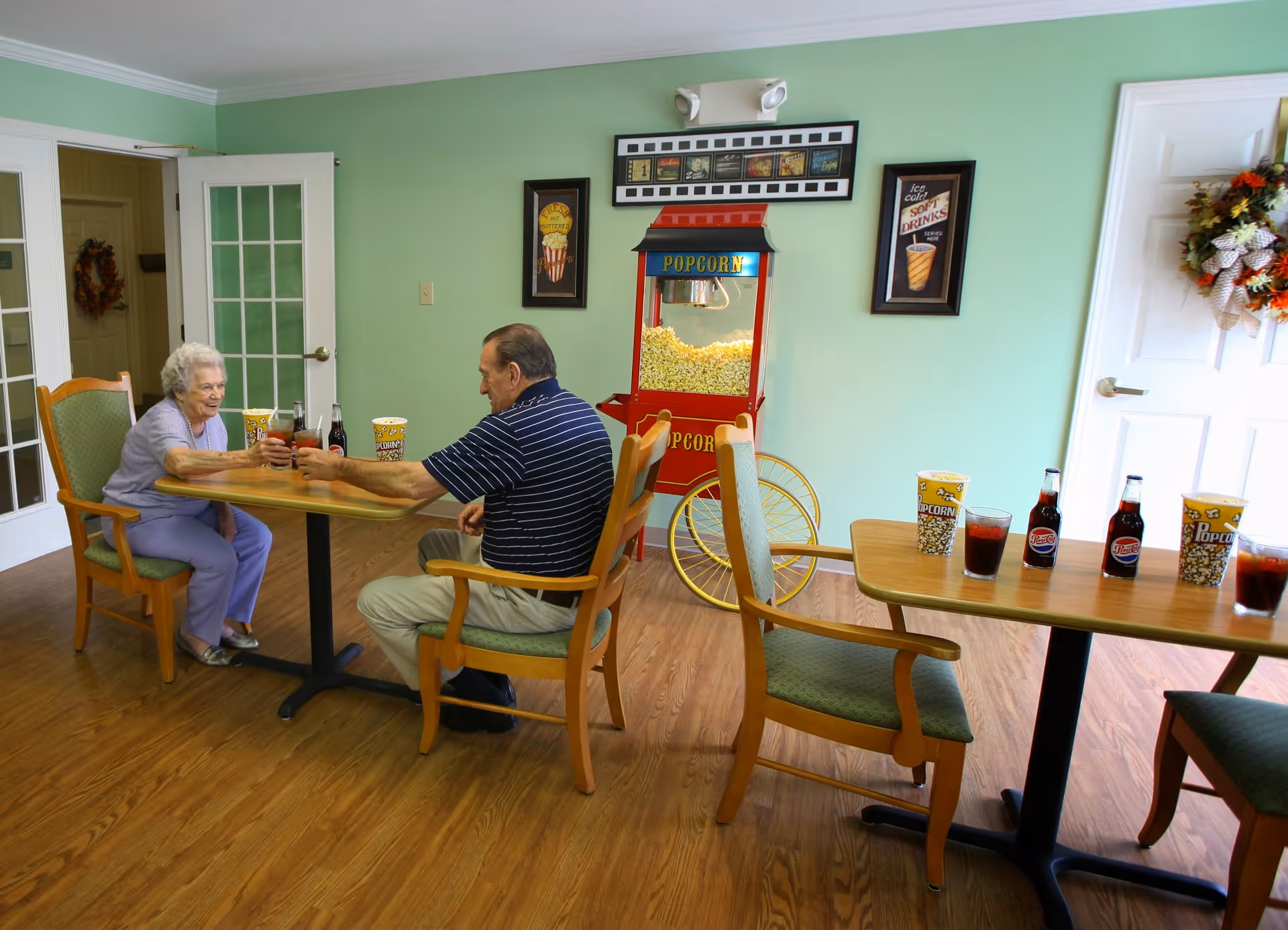 Two elderly people sitting at a wooden table in a room with light green walls, sharing a toast with drinks. There is a popcorn machine filled with popcorn against the wall, framed pictures on the wall, and another table with popcorn containers and bottles of soda.