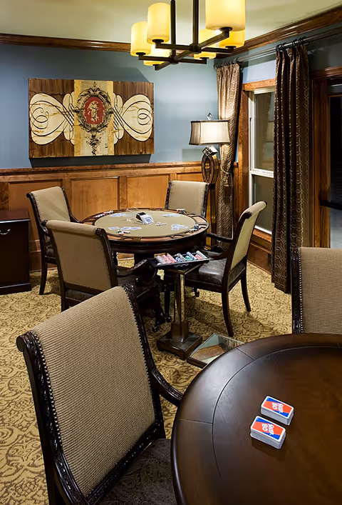 A cozy game room with a round poker table surrounded by four upholstered chairs. The table has playing cards and poker chips on it. Another round table with two decks of cards is in the foreground. The room features patterned carpet, wood paneling on the walls, a floor lamp, and a modern chandelier. A decorative painting hangs on the wall, and windows with brown curtains are visible.