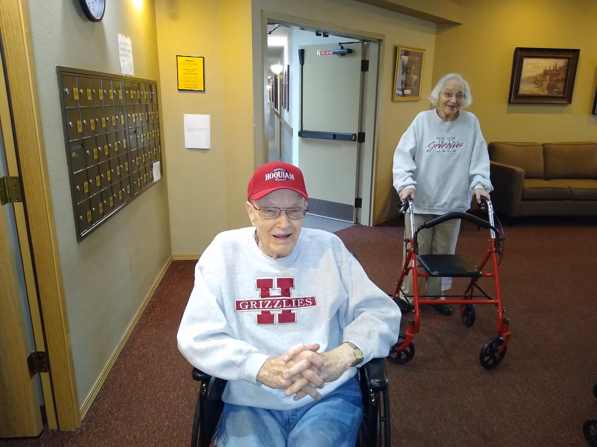 An elderly man in a wheelchair wearing a red cap and a gray sweatshirt with 'Grizzlies' printed on it, smiling. Behind him, an elderly woman using a red walker is also smiling. They are in a hallway with mailboxes on the wall and a brown couch in the background.