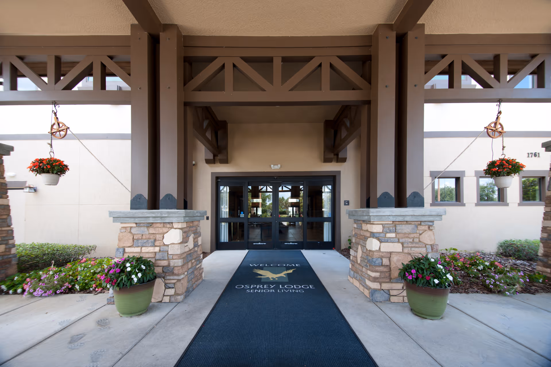 Entrance to a senior living facility with a covered walkway supported by brown beams and stone pillars. There are hanging flower pots with red flowers on either side and potted plants with colorful flowers at the base of the pillars. A black welcome mat with the text 'WELCOME OSPREY LODGE SENIOR LIVING' is placed in front of glass double doors.