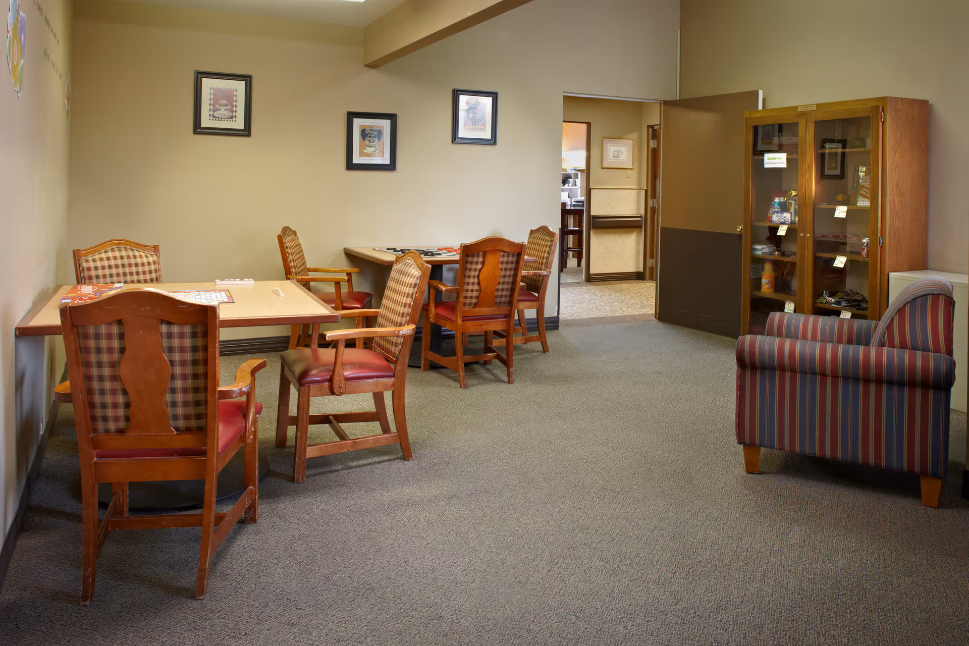 A cozy common area in a senior living facility featuring two wooden tables with several wooden chairs that have plaid upholstery and red cushions. On the right side, there is a striped armchair and a wooden cabinet with glass doors displaying various items. The walls are beige with three framed pictures hanging. An open doorway leads to another room in the background.