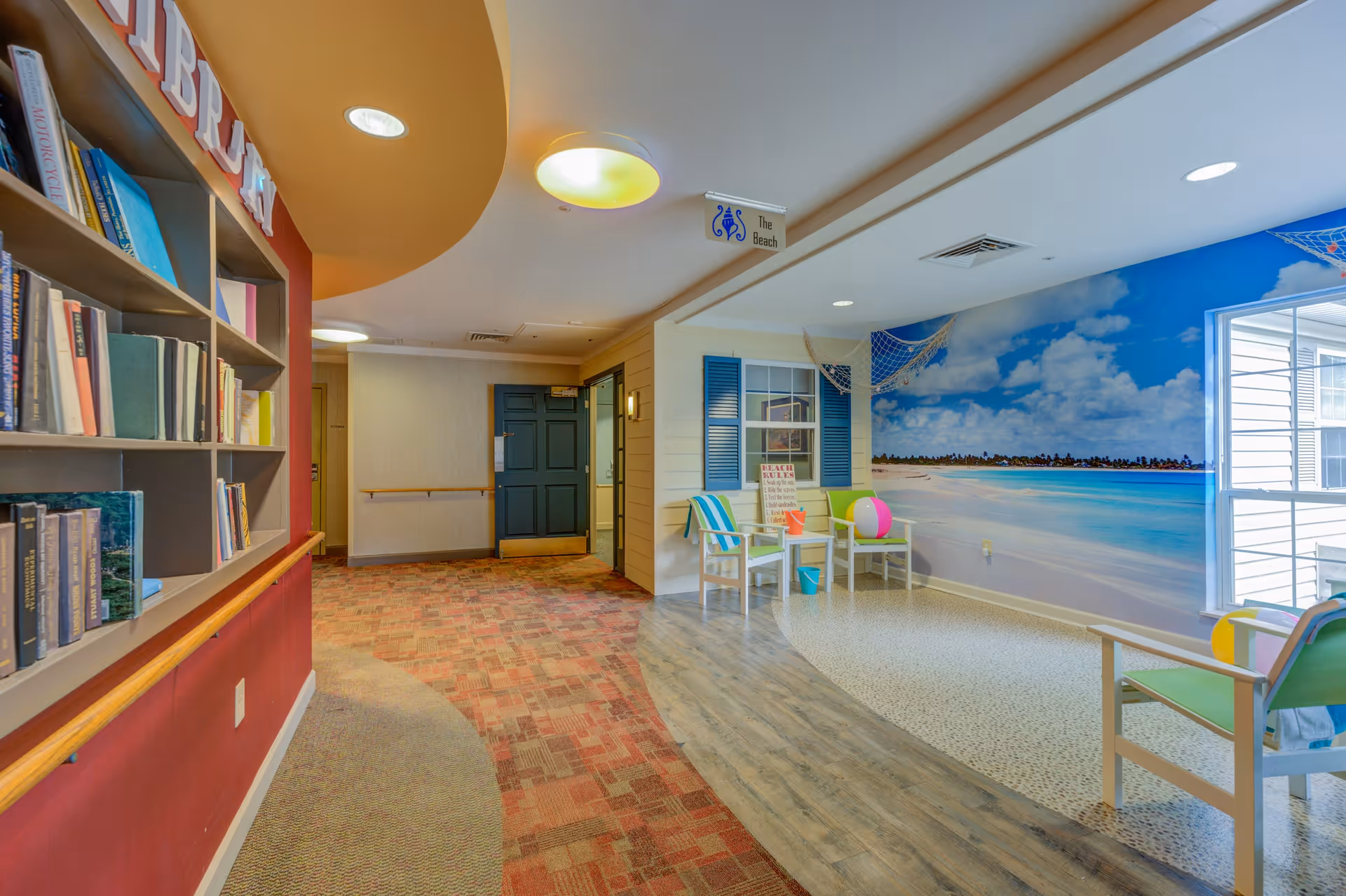 Interior view of a senior living facility hallway with a library area on the left featuring bookshelves filled with books. On the right, there is a beach-themed seating area with chairs, beach balls, and a large mural of a beach with blue skies and ocean. The floor transitions from carpet to wood and tile, and there is a sign hanging from the ceiling that says 'The Beach'.