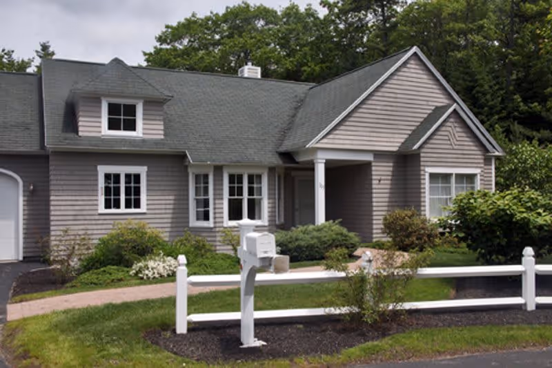Exterior front view of a single-story house with gray siding, white trim, a green shingled roof, a white mailbox, and a white fence in a landscaped yard with bushes and trees in the background.