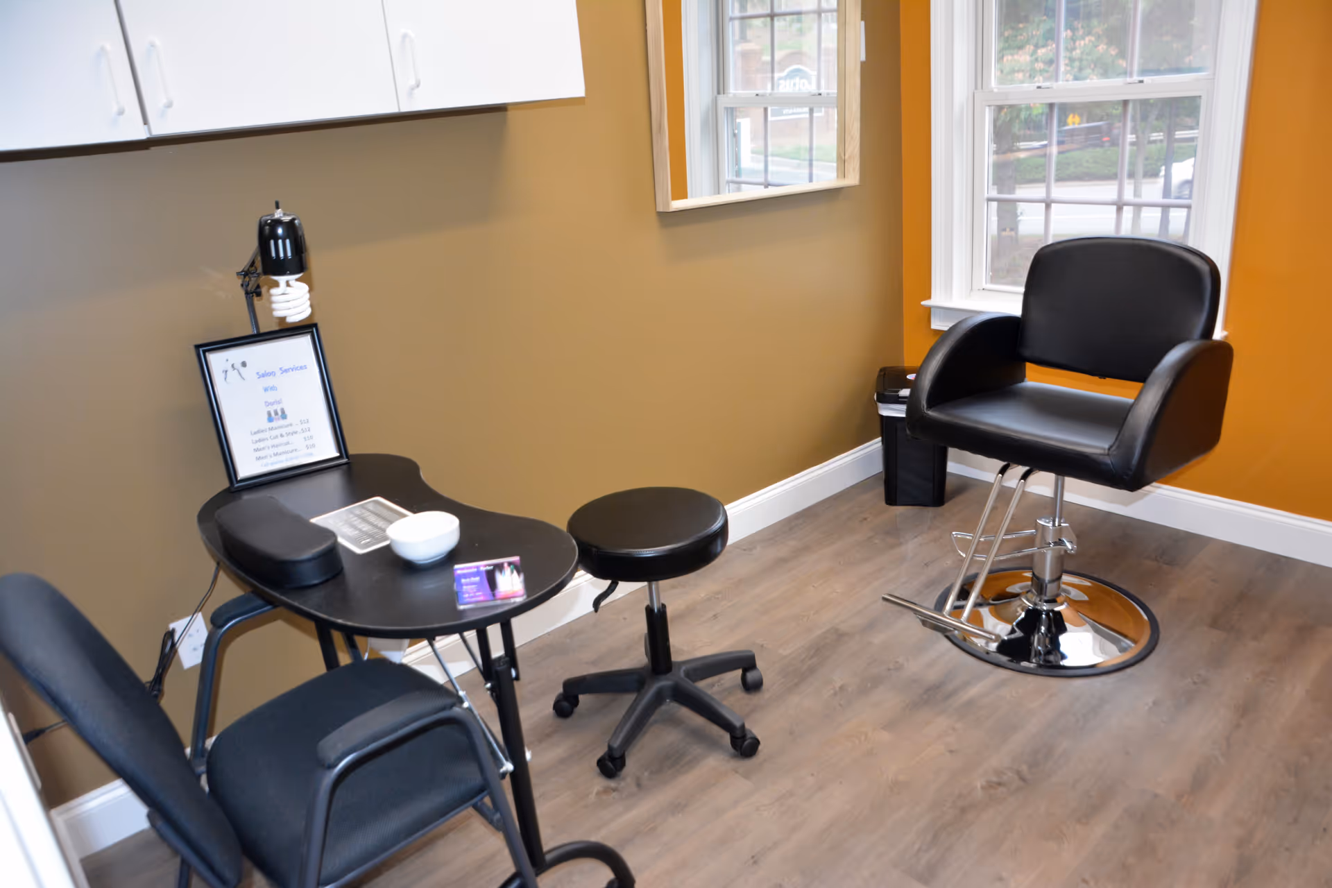 A small salon or grooming area with a black salon chair, a black rolling stool, a black table with a white bowl, a small card, and a framed list of salon services. The room has wood flooring, tan and orange walls, white cabinets, and a window showing an outdoor view.