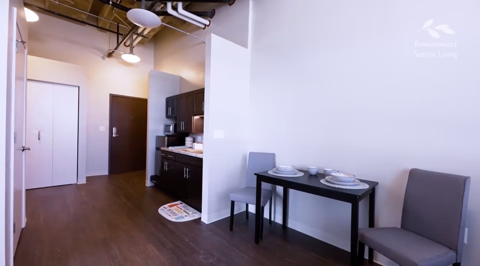 Interior view of a supportive living facility showing a small dining area with a black table set for two with dishes and two gray chairs. To the left is a kitchenette with dark cabinets, a microwave, and countertop items. The floor is dark wood, and the ceiling has exposed pipes and lighting fixtures. A closed door and closet are visible in the background.