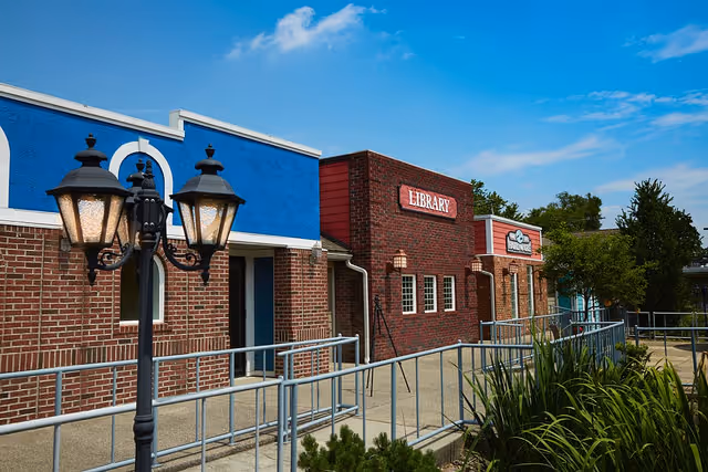 Exterior view of a brick building with a blue and white facade and a red brick section labeled 'LIBRARY'. There is a black street lamp with three lanterns in the foreground, a metal handrail along a concrete walkway, and greenery including bushes and trees. The sky is clear and blue.
