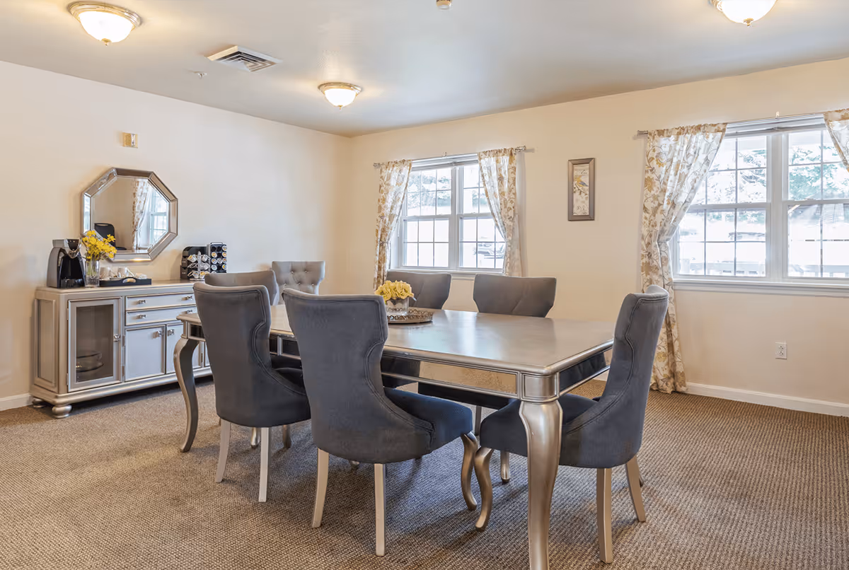 A dining room with a rectangular table surrounded by six upholstered chairs. The room has beige walls, carpeted floor, two windows with floral curtains, and a sideboard with a mirror above it. There is a coffee maker and a flower vase on the sideboard.