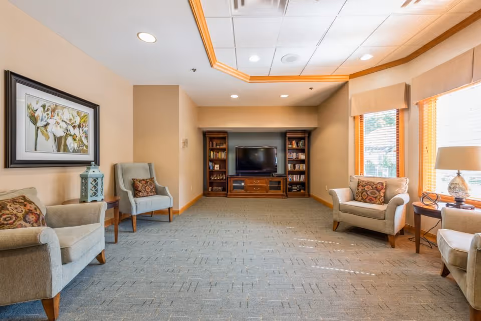 Bright senior living lounge with upholstered armchairs, a TV and bookcases at the far wall, and large windows on the right.