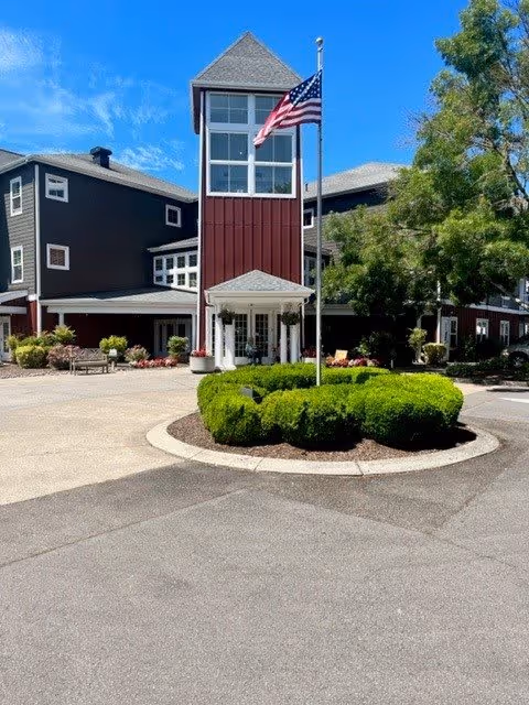 Exterior view of a senior living facility building with a tall central tower featuring large windows. The building is painted in dark red and gray with white trim. In front of the building is a circular landscaped area with neatly trimmed bushes and an American flag on a flagpole. Trees and a clear blue sky are visible in the background.