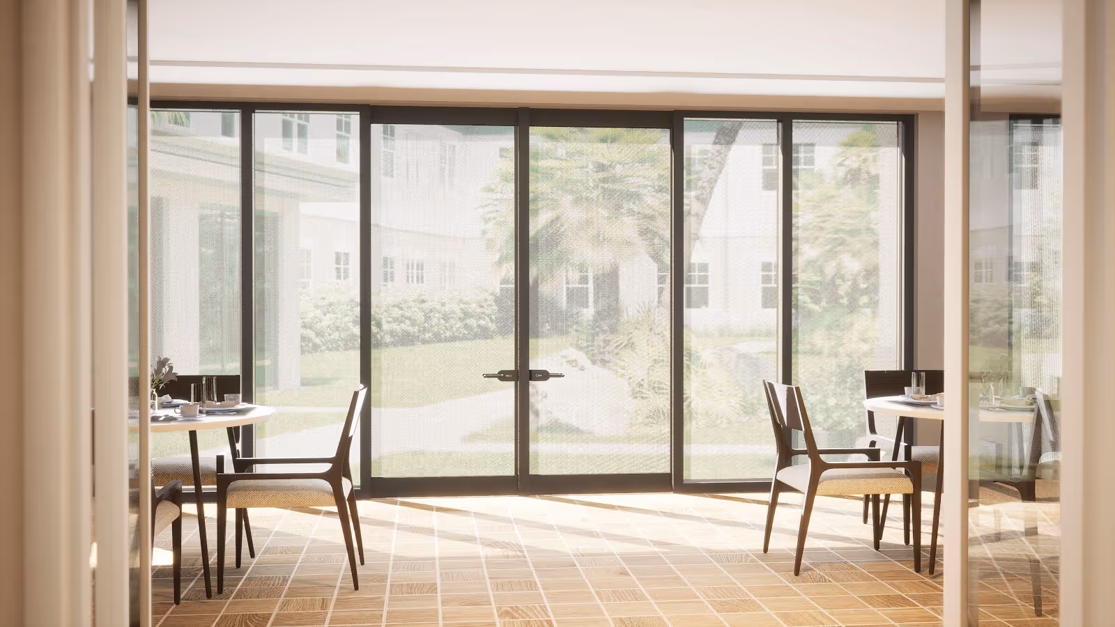 A bright dining area with round tables and chairs arranged near large floor-to-ceiling glass doors that open to a garden outside. Sunlight streams through the glass, illuminating the tiled floor and creating a warm, inviting atmosphere.