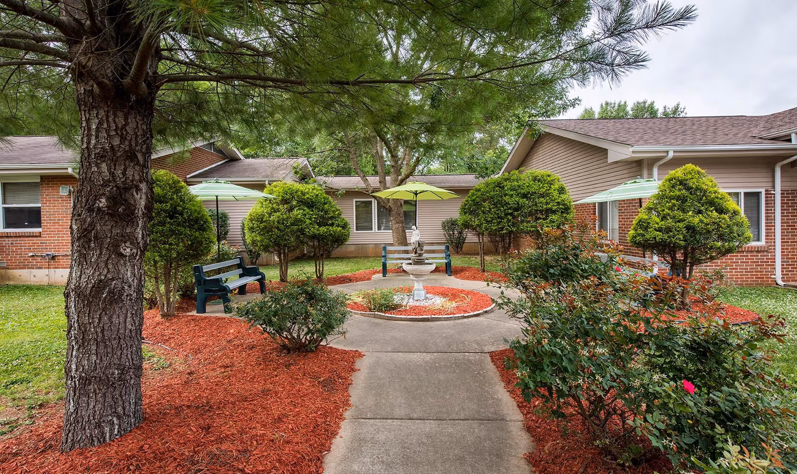 Outdoor courtyard area at Farmington Presbyterian Manor featuring a concrete pathway leading to a circular garden with a small fountain in the center. The garden is surrounded by neatly trimmed bushes, red mulch, and several green umbrellas providing shade over benches. The background shows single-story brick and siding buildings with windows.