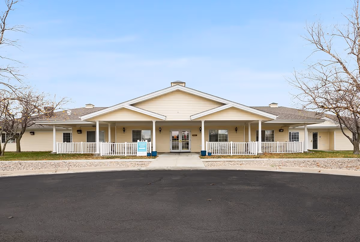 Front exterior view of a single-story senior living facility building with beige siding, a covered entrance with white railings, and a paved driveway in front. Leafless trees are visible on either side of the building under a clear blue sky.