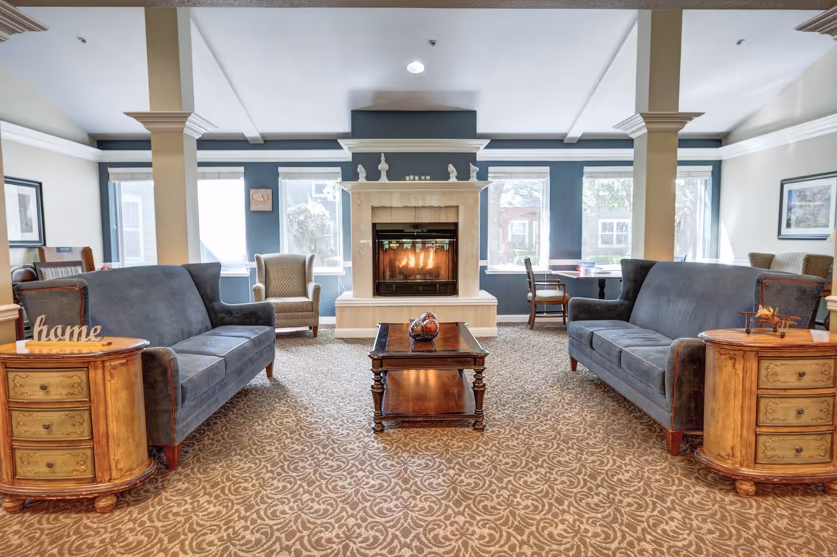Spacious senior living lounge with two blue sofas facing a lit fireplace, a coffee table, and armchairs by large windows.