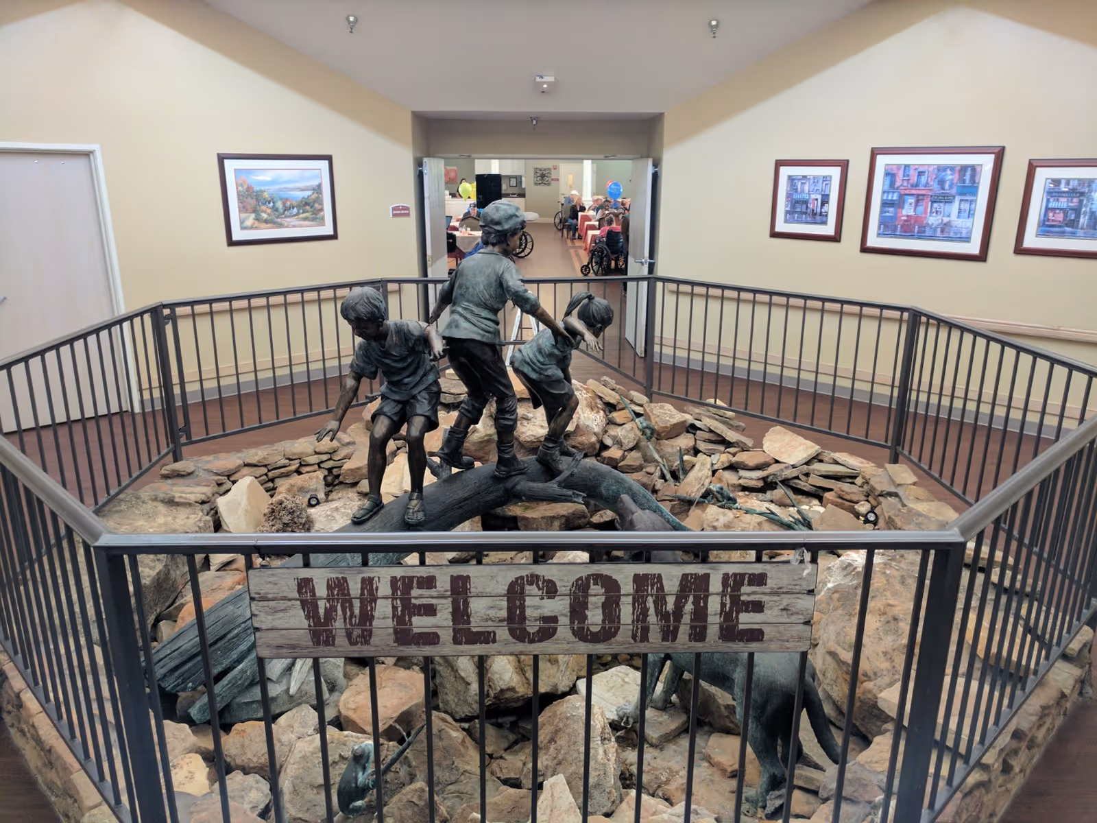 Indoor area of a senior living facility featuring a fenced display with a bronze statue of three children playing on a log surrounded by rocks. A wooden sign with the word 'WELCOME' is attached to the fence. The walls are decorated with framed pictures, and through an open doorway, a dining area with tables and people in wheelchairs is visible.