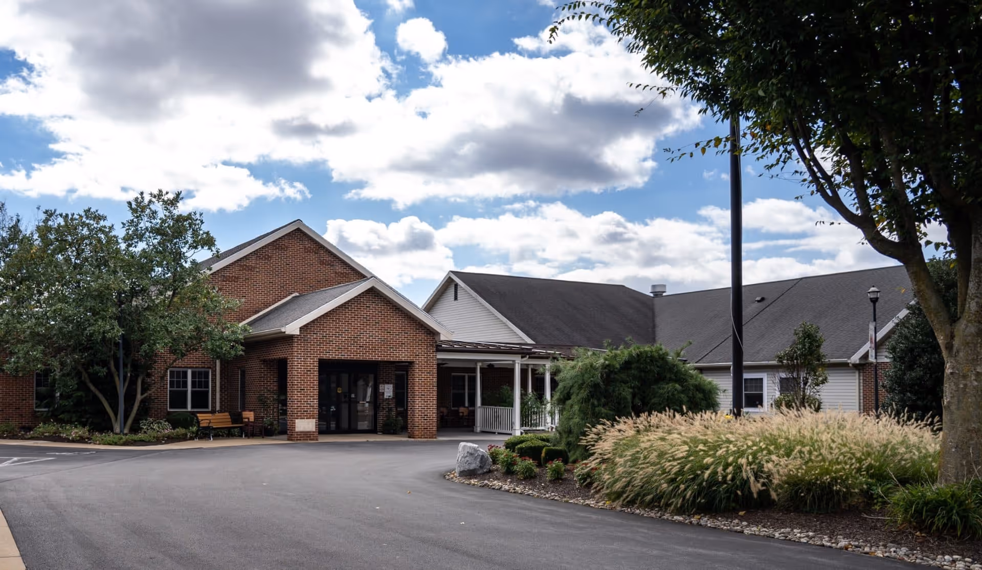 Exterior view of a single-story brick and siding building under a partly cloudy sky. The building has a covered entrance with glass doors, surrounded by landscaped bushes, trees, and ornamental grasses. A paved driveway leads to the entrance.