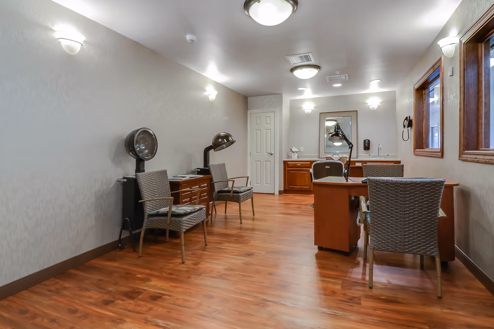 Well-lit interior salon/activity room with wooden floors, desks, salon hair dryers and woven chairs.