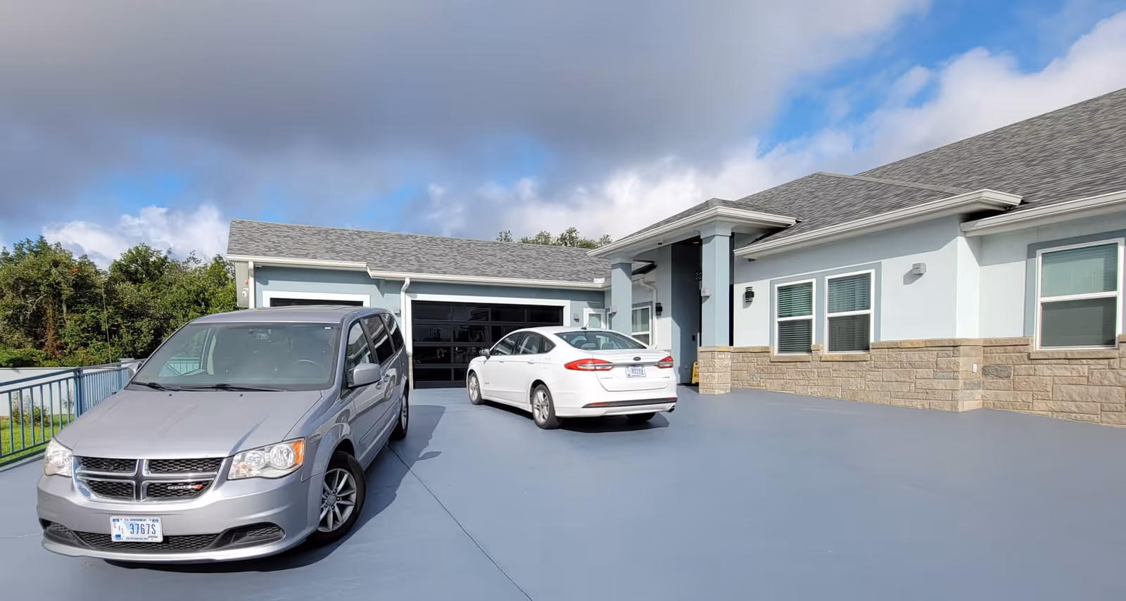 Exterior view of a single-story assisted living facility building with light blue walls and stone accents. Two cars, one silver minivan and one white sedan, are parked on a large paved driveway in front of the building. The sky is partly cloudy.
