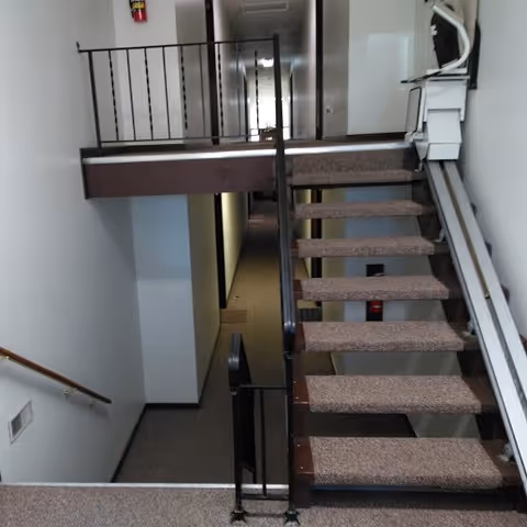 Interior view of a stairwell in Hancock Apartments showing carpeted stairs with metal railings leading to a hallway with multiple doors. The walls are white, and there is a fire extinguisher mounted on the wall at the end of the hallway.