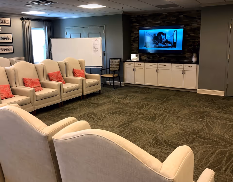 A cozy living room area with beige armchairs arranged in rows facing a wall-mounted flat screen TV. The TV is on, displaying a black and white image. Below the TV is a white cabinet with multiple drawers and doors. To the left, there is a whiteboard on a stand and a single chair. The room has green patterned carpet and light blue walls with framed pictures and a window with curtains.