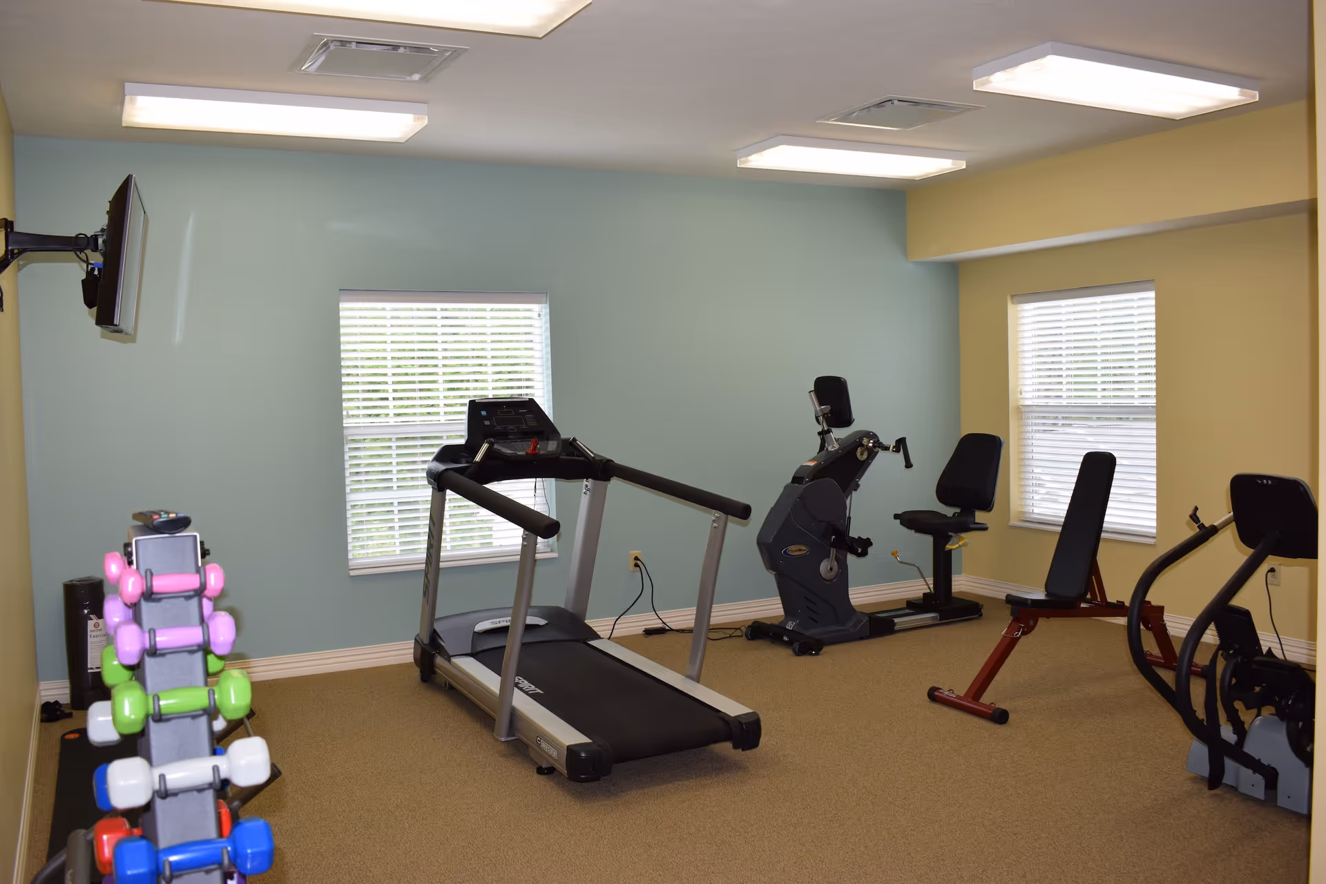 A small fitness room with exercise equipment including a treadmill, stationary bike, weight bench, elliptical machine, and a rack of colorful dumbbells. The room has light blue and yellow walls, two windows with blinds, and ceiling lights.