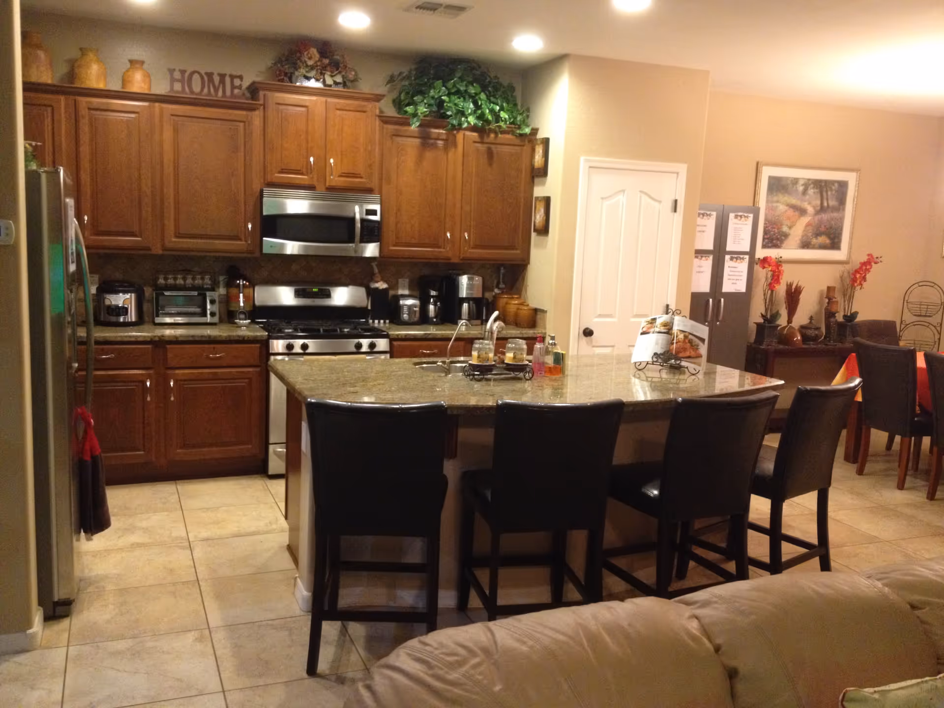 A kitchen area with wooden cabinets, a stainless steel stove and microwave, and a granite countertop island with four black chairs. The kitchen is well-lit with recessed ceiling lights. There are various kitchen appliances on the counter, decorative plants above the cabinets, and a dining area visible in the background with a table, chairs, and wall art.