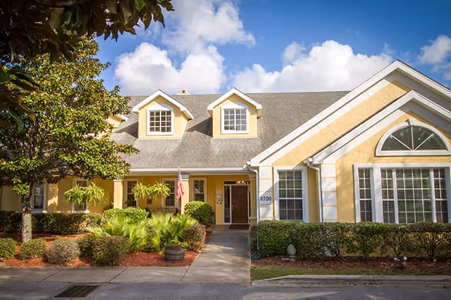 Front exterior of a yellow single-story senior living building with a covered entrance, dormer windows, landscaped shrubs, and an American flag.