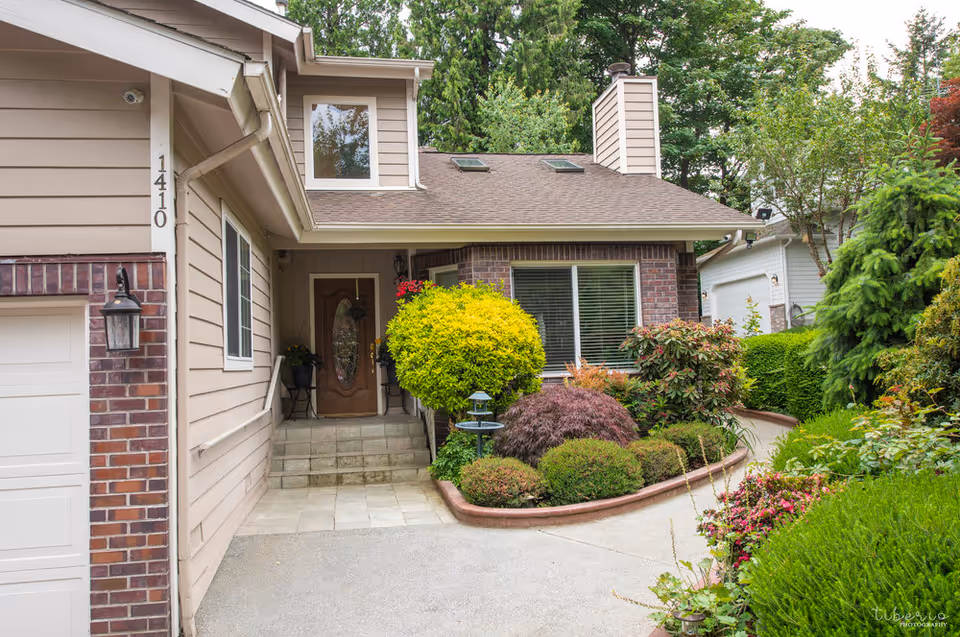 Front exterior view of a residential building with beige siding and brick accents, a wooden front door with glass panel, steps leading up to the entrance, and a well-maintained garden with various shrubs and bushes along a curved pathway.