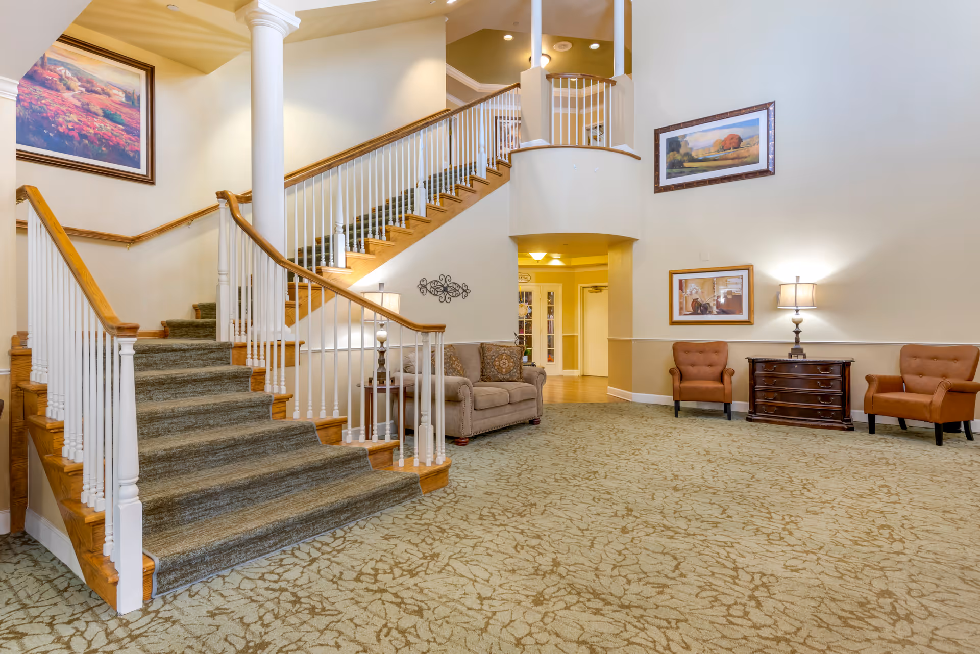 Spacious senior living lobby with a carpeted staircase, seating area, and framed artwork on the walls.