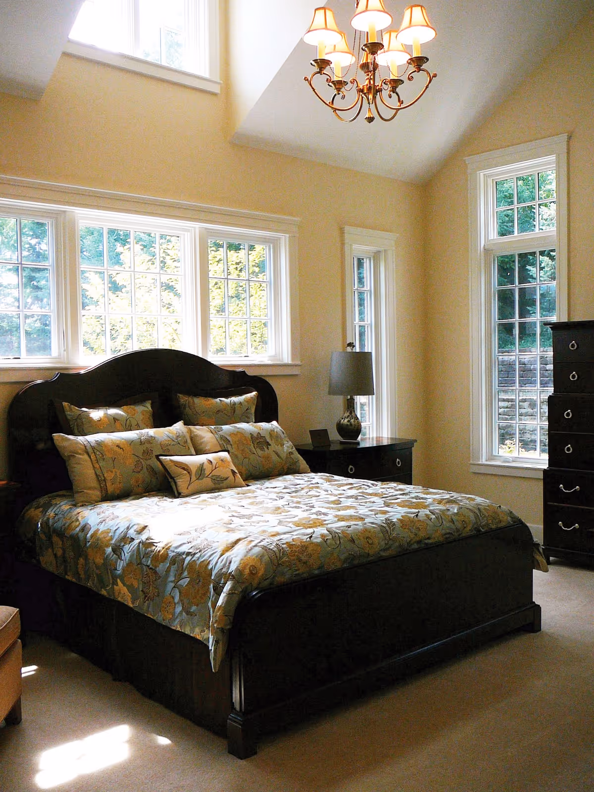 A bright bedroom with a large dark wooden bed featuring floral patterned bedding and multiple pillows. The room has beige walls, several tall windows letting in natural light, a dark wooden nightstand with a lamp, and a matching tall dresser. A chandelier hangs from the ceiling.