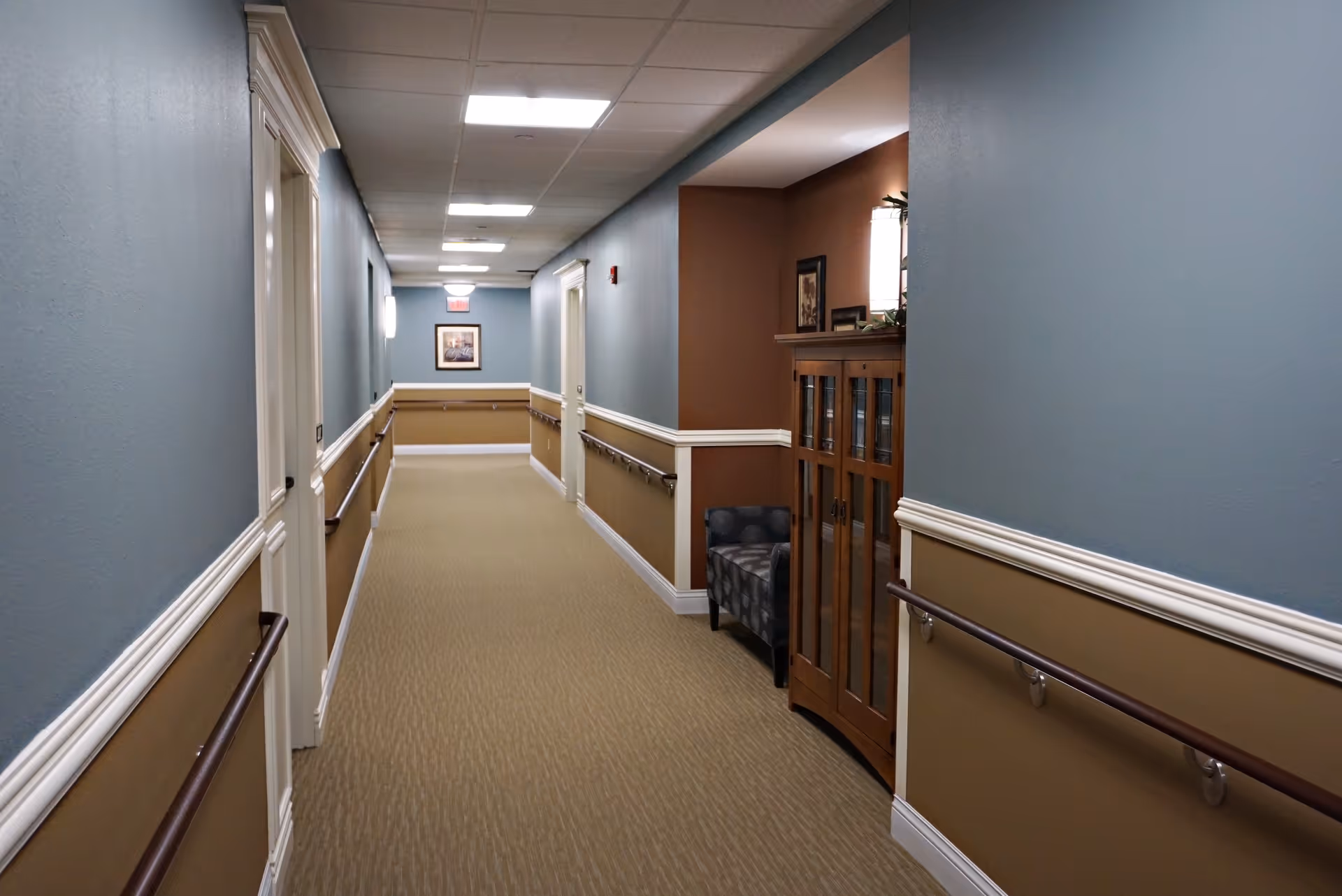 A long, well-lit hallway in an assisted living facility with blue and beige walls, handrails on both sides, a framed picture at the end, and a small seating area with a wooden cabinet and a patterned chair in a recessed nook on the right.