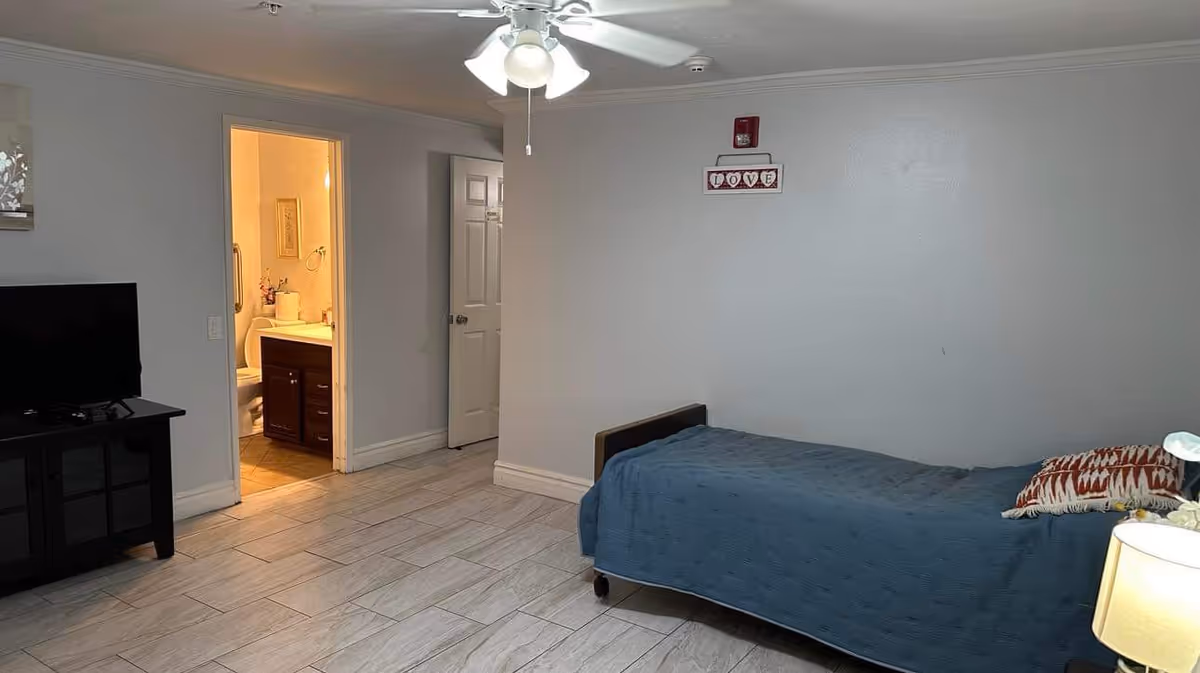 Simple bedroom with a single bed covered by a blue blanket, a TV on a stand, ceiling fan, and an open bathroom door.