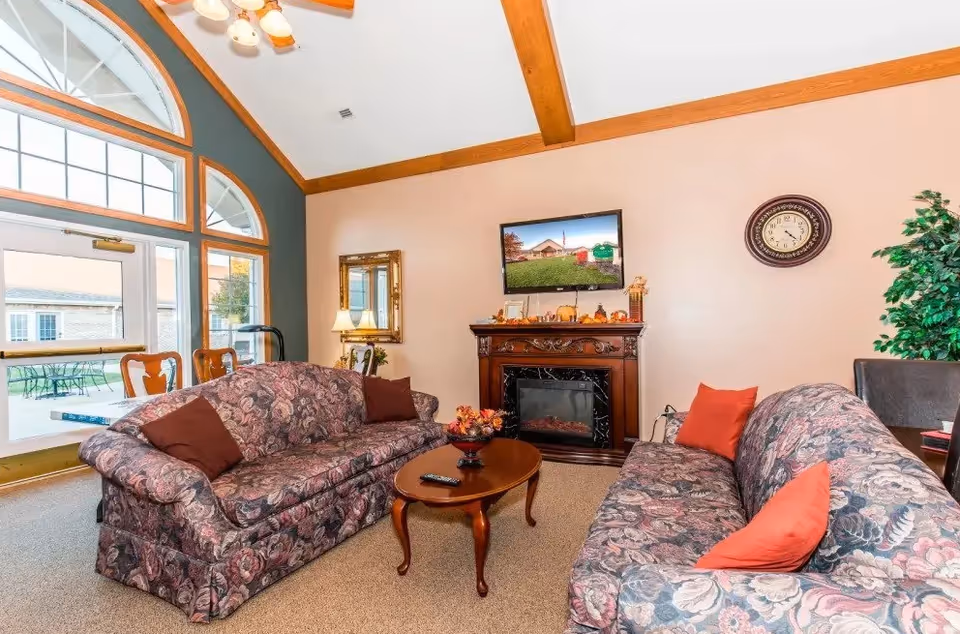 A cozy living room with two floral patterned sofas facing each other, each with colorful throw pillows. A wooden coffee table with a decorative centerpiece and a remote control sits between the sofas. On the wall, there is a flat-screen TV mounted above an ornate electric fireplace decorated with autumn-themed items. A round wall clock and a large green plant are also visible. The room features high ceilings with wooden beams and large windows letting in natural light, with a view of an outdoor patio area.
