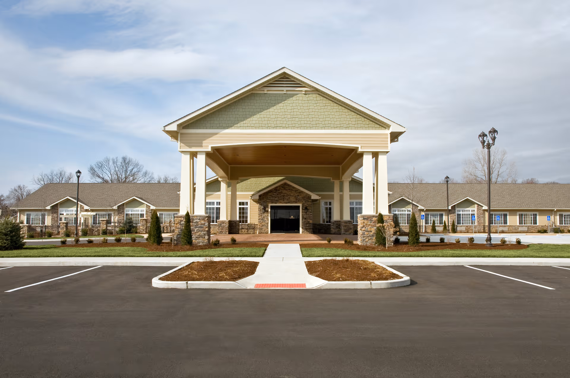 Front exterior view of a single-story senior living facility building with a large covered entrance supported by columns. The building has a combination of stone and siding exterior with multiple windows and a neatly landscaped area in front. The parking lot is empty and the sky is partly cloudy.
