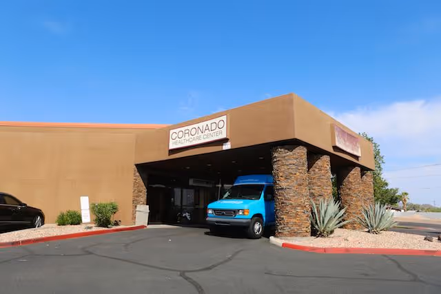 Exterior view of Coronado Healthcare Center showing the entrance with a covered driveway supported by stone pillars. A blue van is parked under the entrance canopy, and there are desert plants and a clear blue sky in the background.