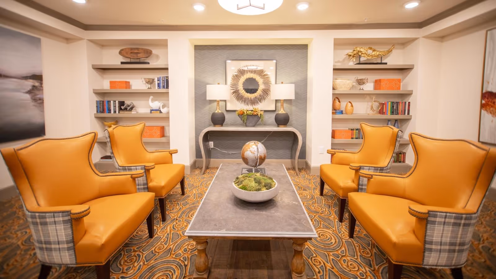 A cozy seating area in an assisted living facility featuring four mustard yellow armchairs with plaid sides arranged around a rectangular coffee table. The table holds a decorative globe and a bowl with greenery. Behind the seating area is a console table with two lamps and a framed artwork, flanked by built-in shelves filled with books and decorative items. The room has a patterned carpet and soft lighting.
