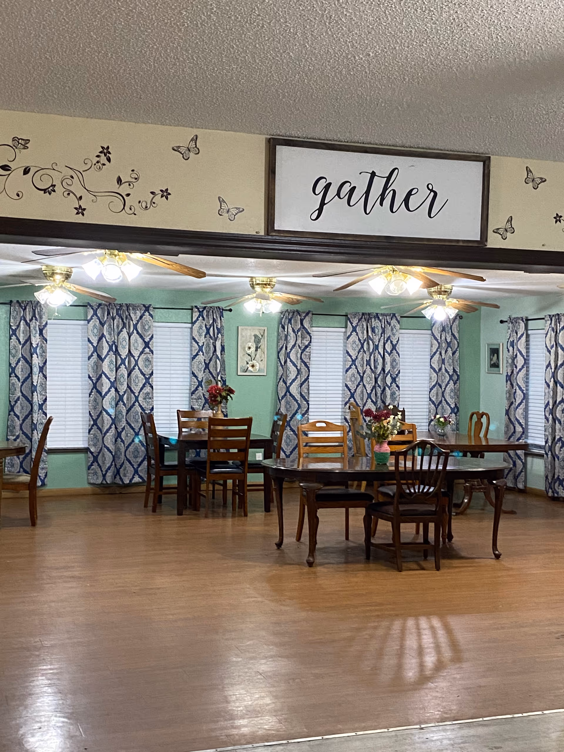 Spacious dining room with wooden tables and chairs, patterned curtains, ceiling fans, and a framed sign reading "gather" above.