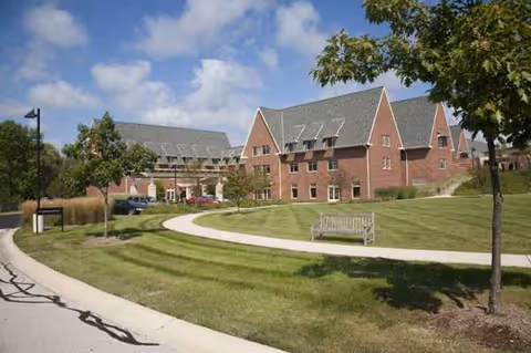Brick senior living campus with curved walkway, manicured lawn, trees, and a bench.