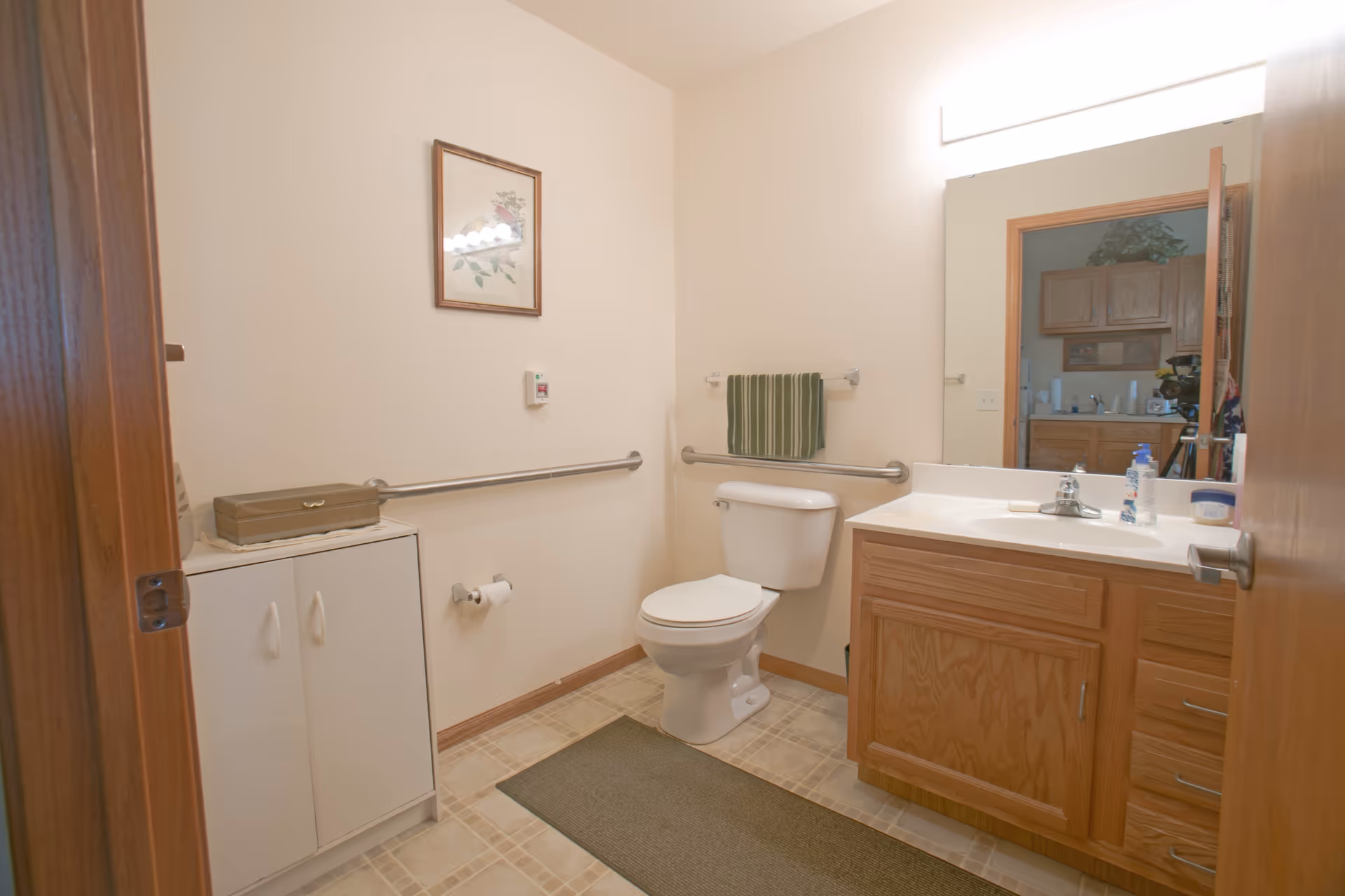 A clean bathroom with a white toilet, wooden vanity with a sink, a large mirror above the sink, a towel rack with a green striped towel, a small cabinet, and a framed floral picture on the wall. The floor has light-colored tiles and a green rug in front of the toilet.