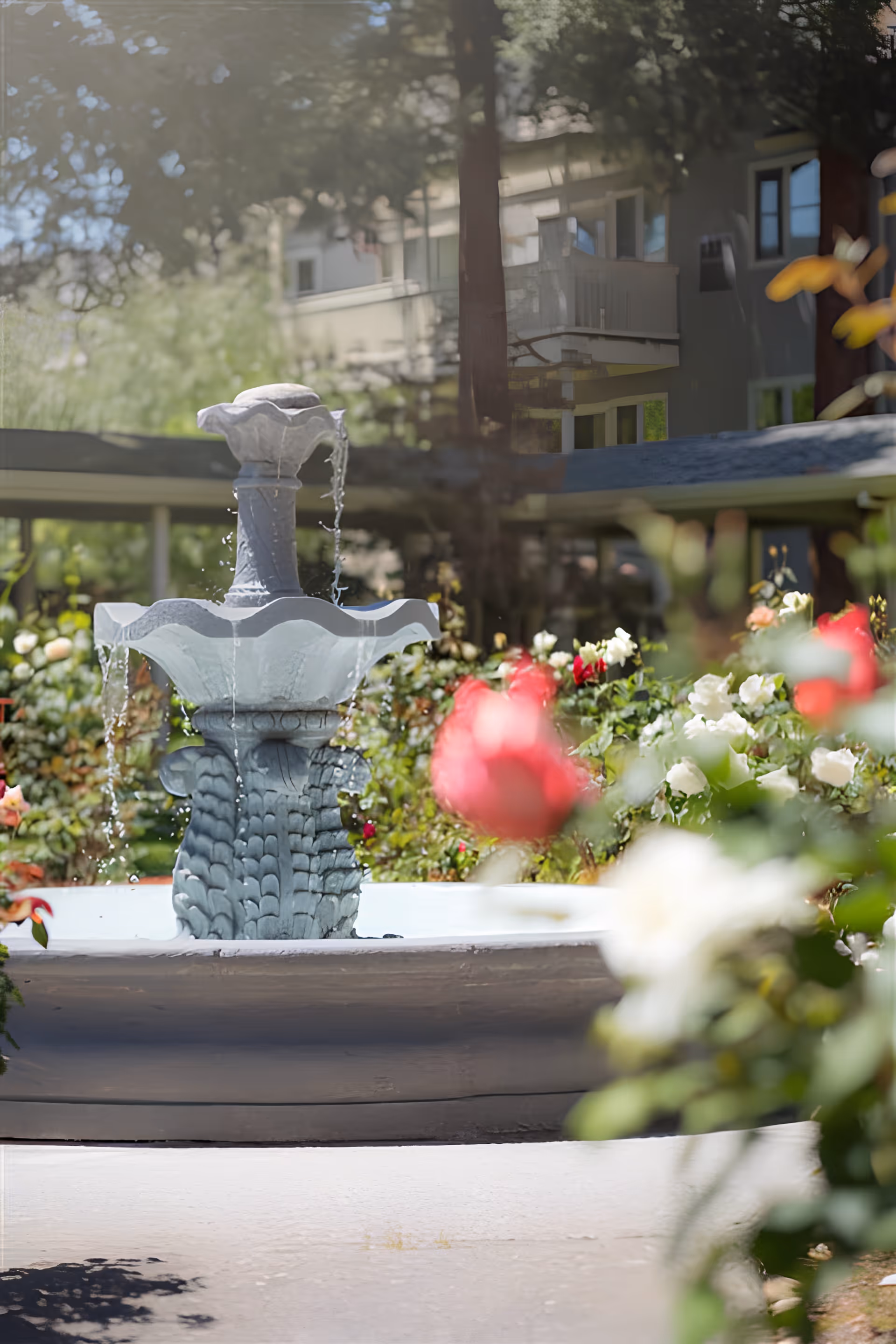 A decorative water fountain in the center of a garden with blooming red, white, and pink flowers. In the background, there is a building with balconies partially visible through the trees.