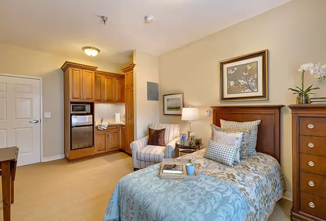 A cozy bedroom in a senior living facility featuring a single bed with multiple pillows and a blue patterned bedspread. Next to the bed is a wooden nightstand with a lamp, a small framed photo, and a plant. A comfortable armchair with a brown pillow is placed beside the nightstand. The room also includes a small kitchenette with wooden cabinets, a microwave, and a mini refrigerator. The walls are painted beige, and there are two framed pictures hanging on the walls.