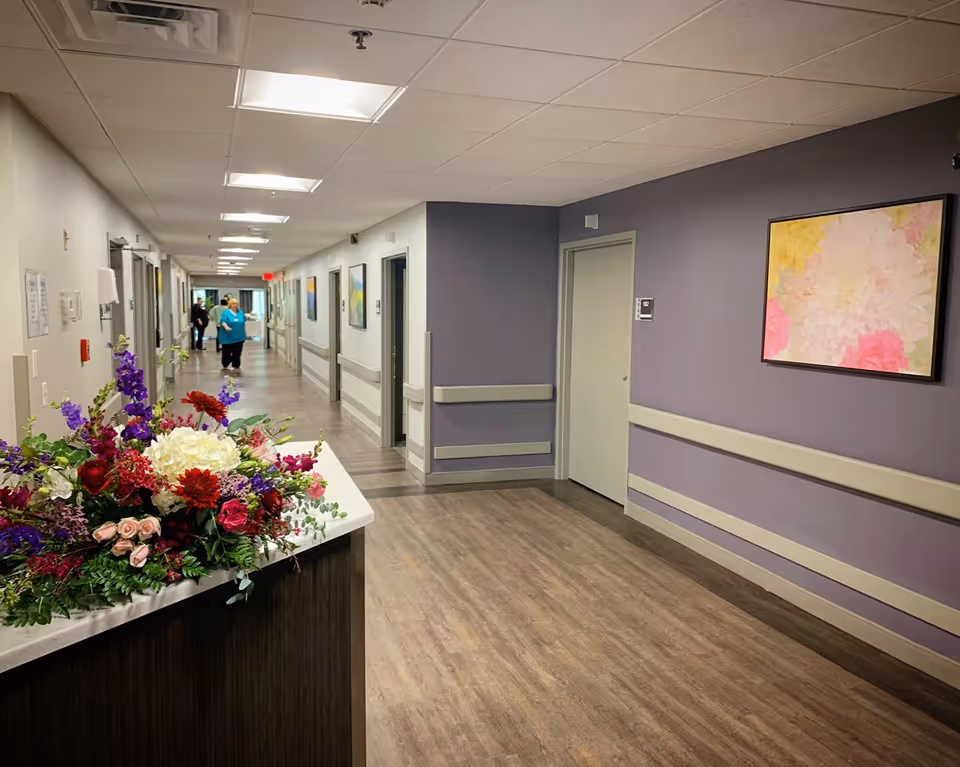 Long interior hallway of a senior living facility with a floral arrangement on a reception counter, purple walls, handrails and staff walking in the distance.