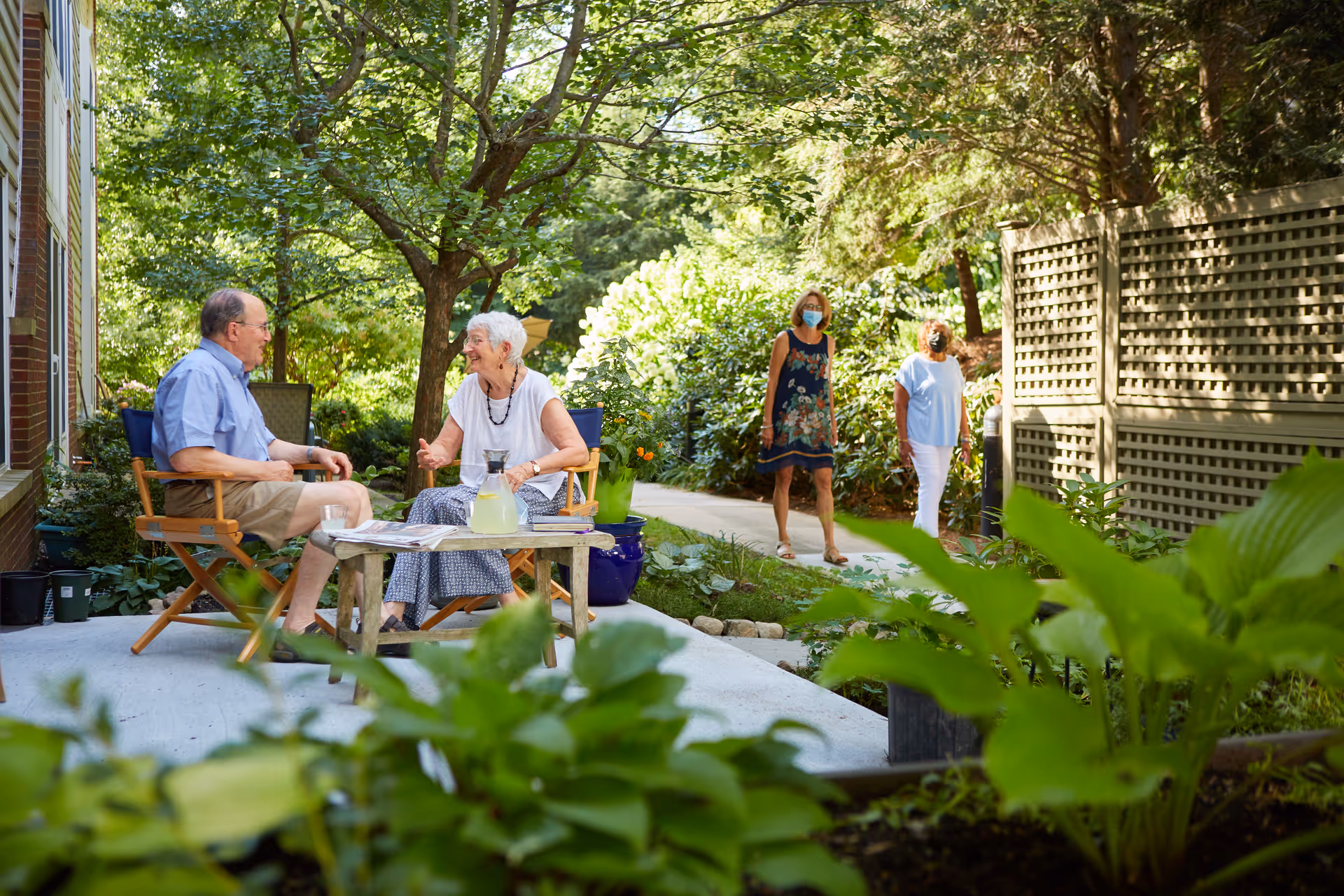 Two elderly people sitting and talking on wooden chairs at a small table with a pitcher of lemonade outside in a garden area, while two women wearing masks walk on a path nearby surrounded by lush greenery and trees.