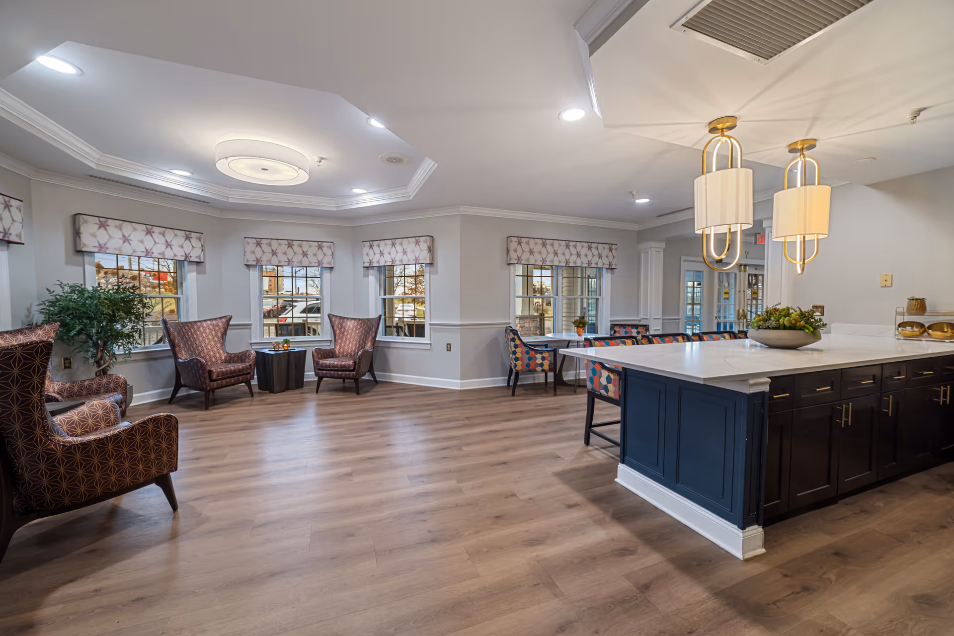 Open, well-lit senior living lounge with armchairs by windows and a large kitchen island with pendant lights.