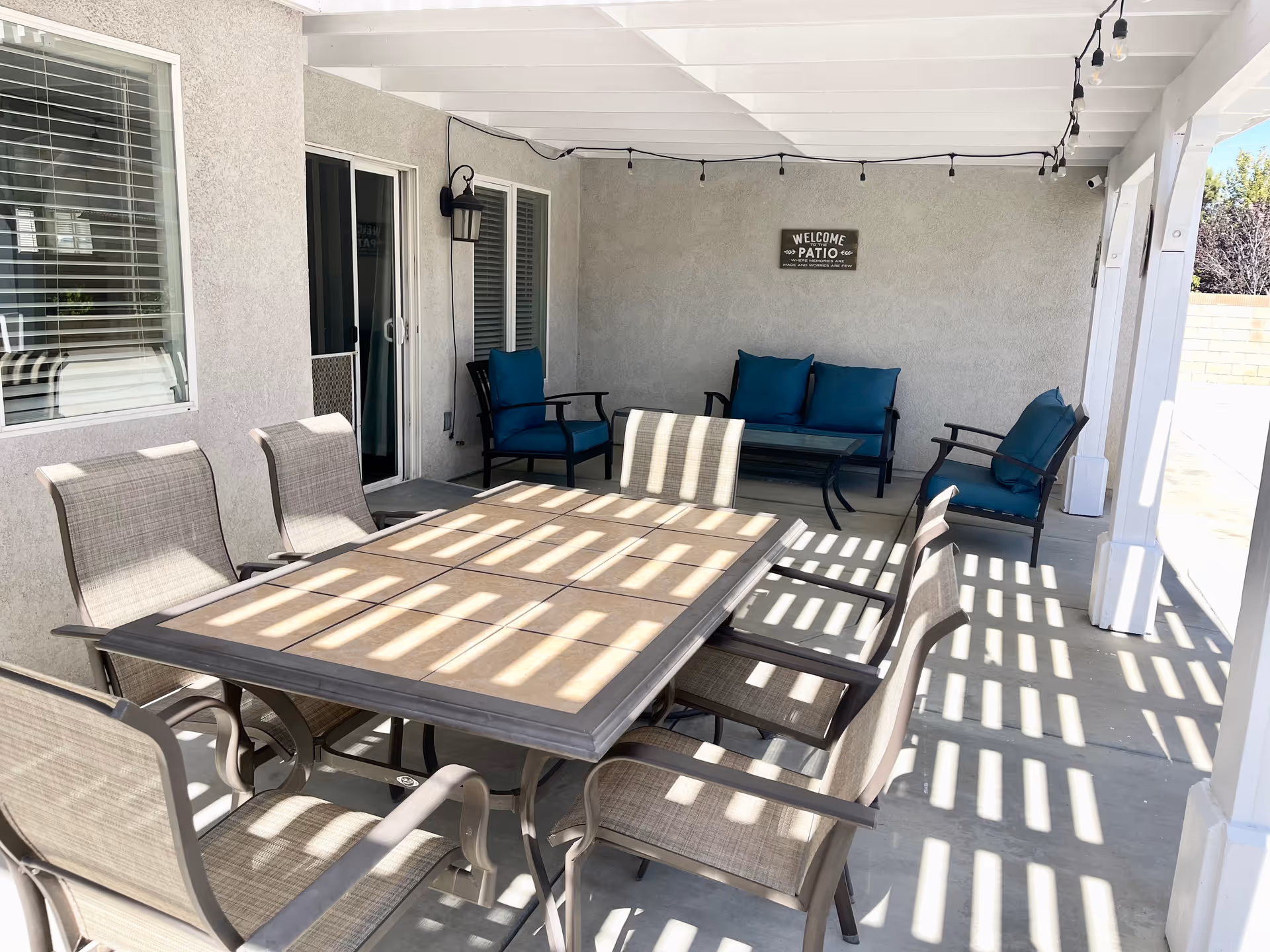 Covered outdoor patio area with a large rectangular table surrounded by eight chairs. In the background, there are three cushioned armchairs and a cushioned loveseat with a glass coffee table. String lights hang from the ceiling, and a sign on the wall reads 'Welcome Patio'.