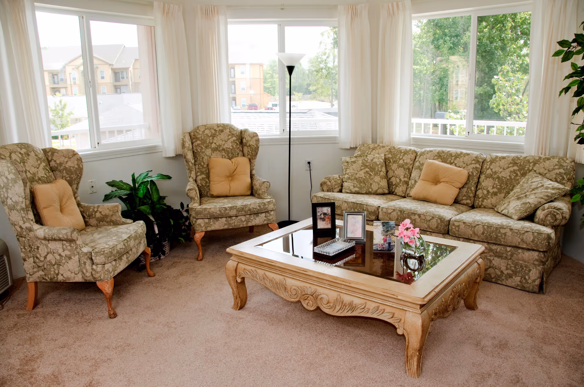 A bright living room with large windows showing an outside view of buildings and trees. The room features a floral patterned sofa and two matching armchairs with beige cushions. A wooden coffee table with a glass top holds framed photos and a small vase with pink flowers. A tall floor lamp stands near the windows, and there are green plants in the corners.