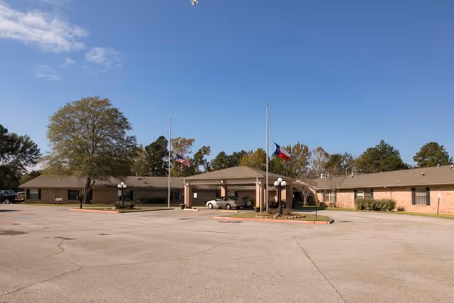 Exterior view of a single-story brick building with a covered entrance and three flagpoles displaying the American flag, the Texas state flag, and another flag. The building is surrounded by trees and a large paved parking area under a clear blue sky.
