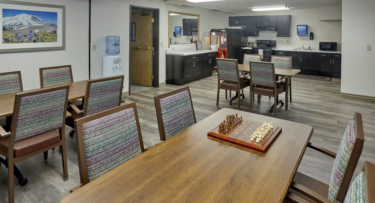 A communal room with multiple wooden tables and chairs with patterned upholstery. One table in the foreground has a chessboard with chess pieces set up. The background shows a kitchenette area with dark cabinets, a microwave, a stove, a popcorn machine, and a water cooler. A framed picture of a mountain landscape hangs on the wall.