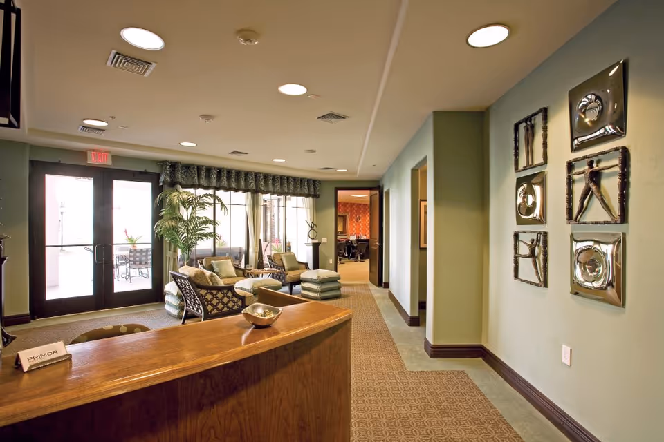 A bright reception lounge with a wooden front desk, seating area, potted plants and glass double doors.