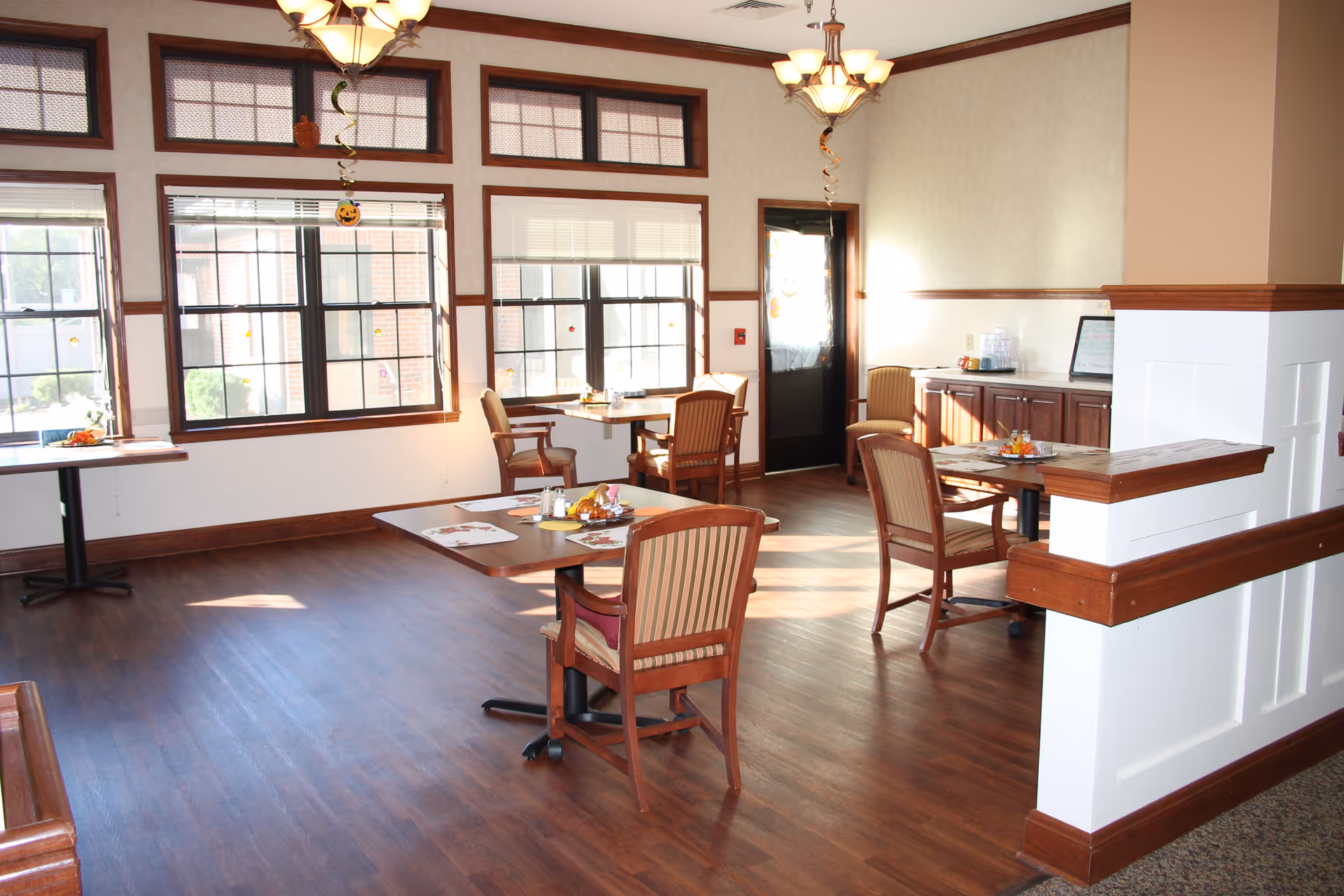 Sunlit dining room with wooden tables and chairs, large windows, and hardwood floors.